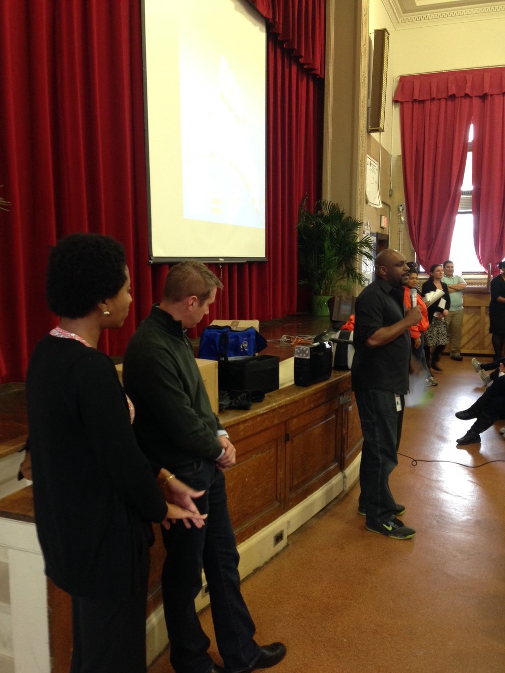 A staff member of DDOE's Energy Patrol unit presents to a packed auditorium of students.  On the left, mentors Mia Dancy and Sierra Dennis ponder Energy Jeopardy questions. 